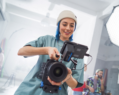 Young Caucasian Female Filmmaker In A Beanie Adjusts A Camera On A Busy Film Set, Capturing The Essence Of Modern Cinematography With Her Team In The Background, Focused On A Creative Project.